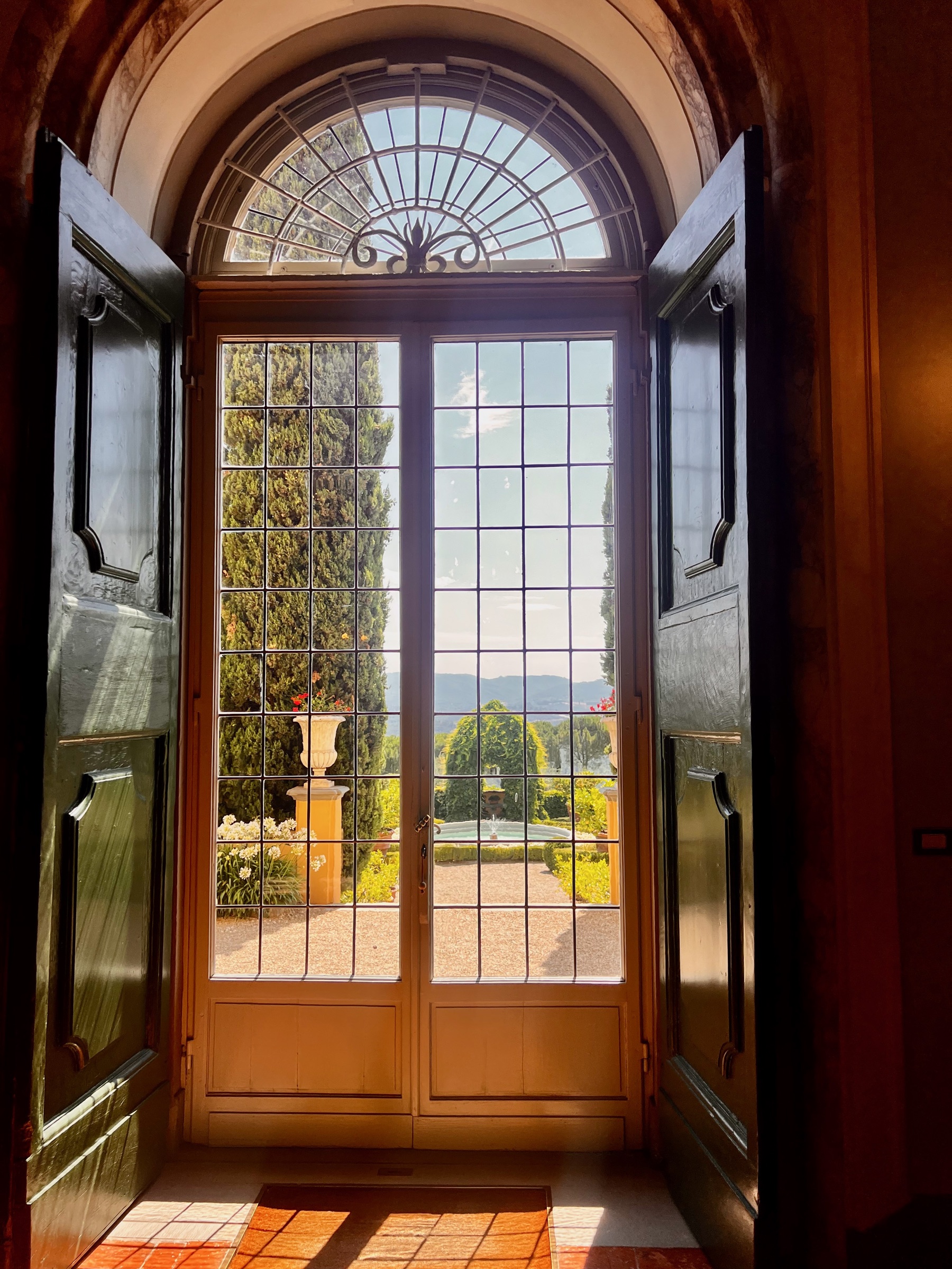 A formal Tuscan garden and distant hills framed by tall glass villa doors